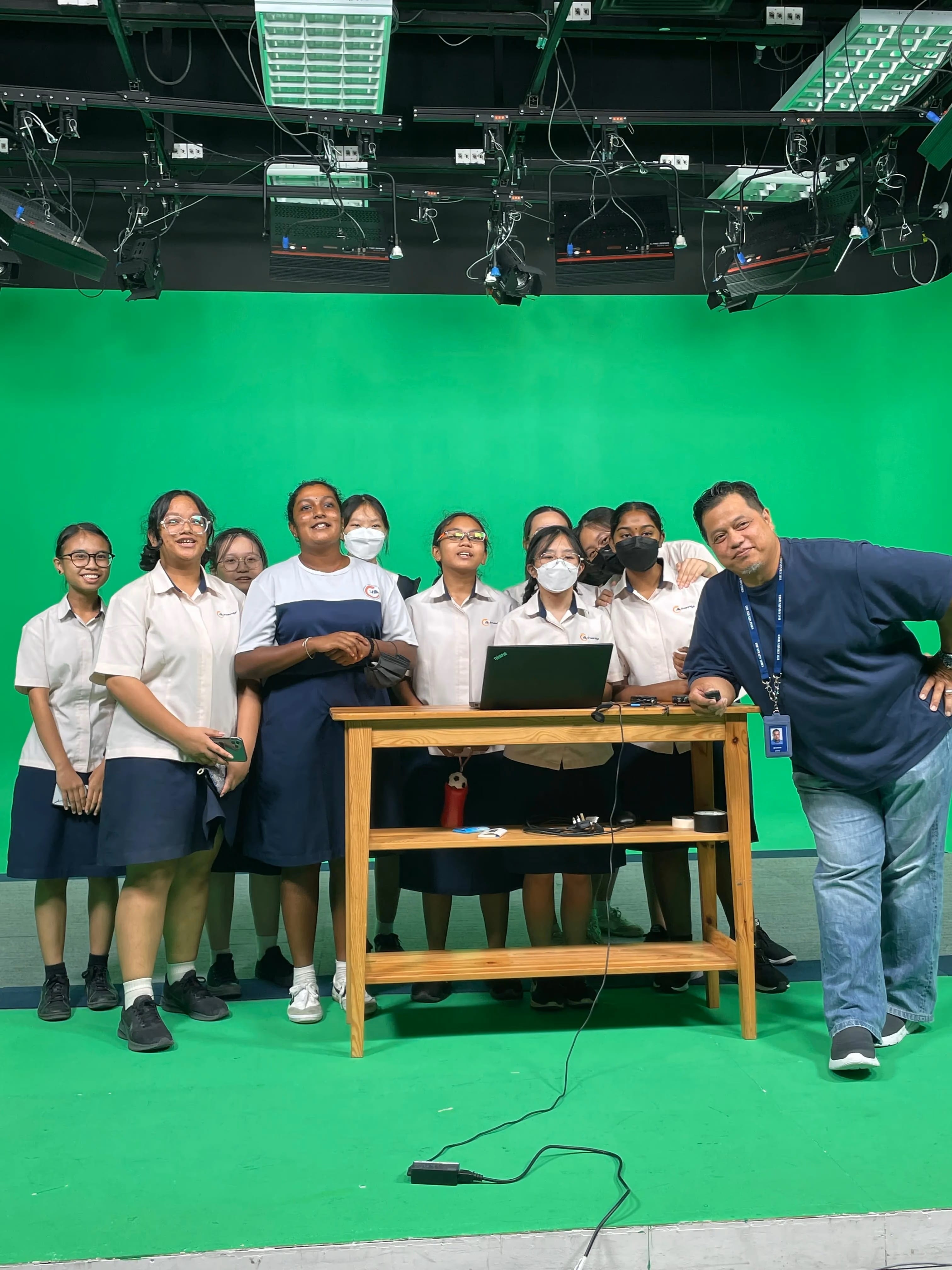 A group of uniformed students and an adult man pose together in a green screen studio with studio lights overhead.